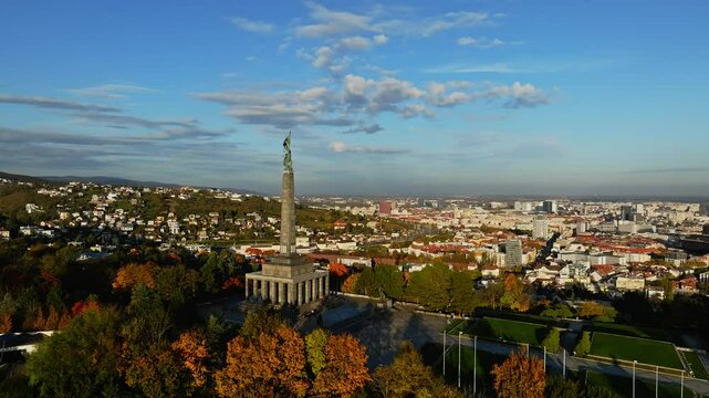 Aerial view over Bratislava showcasing the Slavin War Memorial amidst vibrant autumn colors. The city&rsquo;s urban landscape extends under a clear blue sky, highlighting historic and modern architecture.