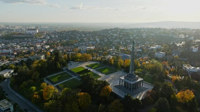 Aerial view over Bratislava, capturing the Slavin War Memorial surrounded by autumn trees. The cityscape stretches out with Bratislava Castle in the distance under a clear sky. (4K)