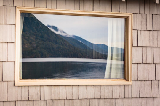 A cabin window reflecting mountains and lake water. 