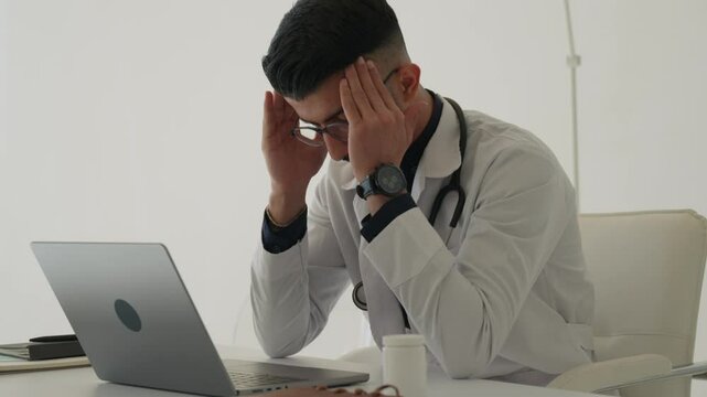 Young hispanic man doctor stressed at clinic or hospital. An overworked young indian male doctor in a white coat expresses exhaustion. Middle Eastern guy therapist having headache, struggling working