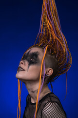 Young woman has an orange colored braided hairdo and horror black stage make-up on face, looking up mysteriously in way that is both captivating and intriguing. Dramatic portrait on blue background