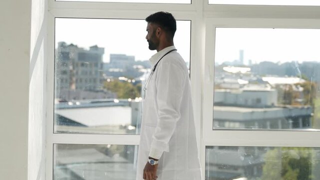 desperate or fatigued indian male therapist in white uniform stands near window feels concerned, looks terribly upset due to medical error, overloaded. Professional failure