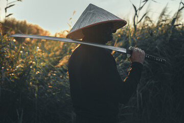 A samurai warrior in a conical hat and black clothes holds a katana on his shoulder at sunset in a field of tall grass. A handsome samurai looks into the distance with a katana on his shoulder.