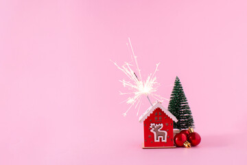 Small red house with glowing sparkler, tree and decoration balls on a pstel pink background. Christmas holiday decoration, a minimal concept with copy space.