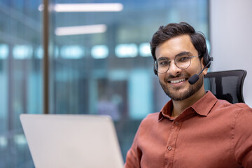 Hispanic businessman wearing headset working on laptop in office setting. Confident smile, brown shirt, glasses, engaging with professionalism. Bright, modern background. Business and communication.