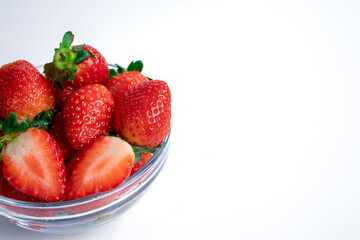 A glass bowl filled with fresh strawberries, some whole and one cut in half, showing vibrant red color and juicy texture on a white background. Ideal for health, nutrition, and food photography themes