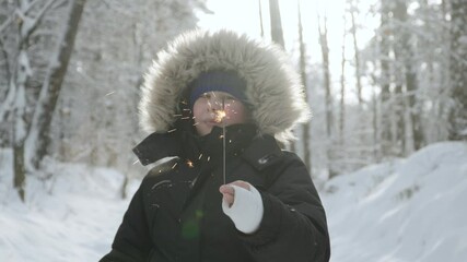Caucasian boy holds Bengal lighted flame in hand in plaster. Portrait of child with injury in frosty snowy park among trees, with smile and festive mood. Optimism and recovery on walk in forest.