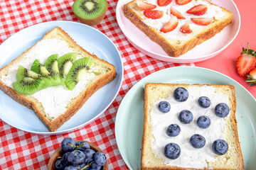 Assorted fruit toasts with blueberries, strawberries, and kiwi on white bread served on colorful plates over a red gingham cloth, with sliced bread, whole fruits, and books in the background