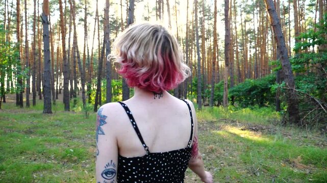 Carefree hippie woman with pink hair having fun at summer forest. Happy smiling punk girl with tattoos fooling around woodland. Young female hipster with ear tunnels enjoying rest at summer nature