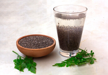 A glass filled with soaked chia seeds alongside a bowl of dry chia seeds, with moringa leaves scattered on the ground, highlighting the nutritious qualities of these superfoods.