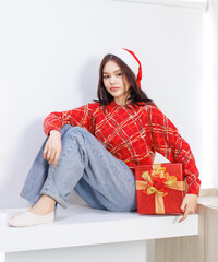 Happy Asian beautiful woman wearing santa hat red long sleeve knitted sweater sitting holding present gift box funny smiling behind white background, Christmas Eve Happy New Year's Party celebration.