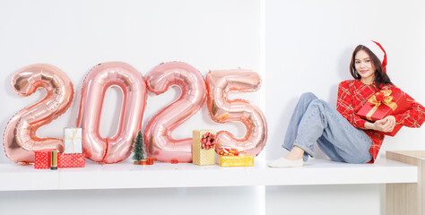 Happy Asian beautiful woman wearing santa hat red long sleeve sitting New year 2025 rose gold balloons on white background with festive decorations ornaments and gift box celebrations holiday concept.