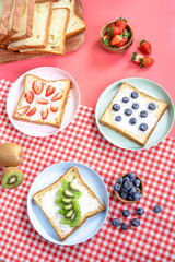 Assorted fruit toasts with blueberries, strawberries, and kiwi on white bread served on colorful plates over a red gingham cloth, with sliced bread, whole fruits, and books in the background