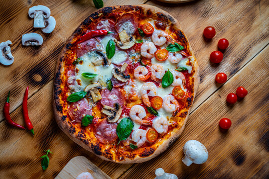 Pizza with two types of toppings, one half paperoni with mushrooms, the other tomatoes and shrimps with cheese. Pizza on a wooden table with ingredients.