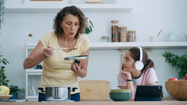 Video of beautiful mother cooking and letting her daughter taste the food while daughter is using digital tablet in the kitchen at home.