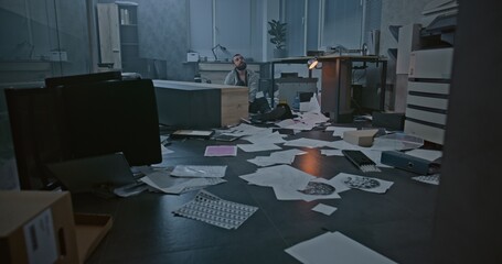 Dolly shot of depressed office worker sitting on the floor in destroyed office room. Frustrated...