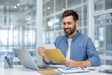 Fototapeta premium Man happily opening envelope at desk with laptop in modern office, illustrating positive emotion, communication, and work success. Bright setting with stylish decor.