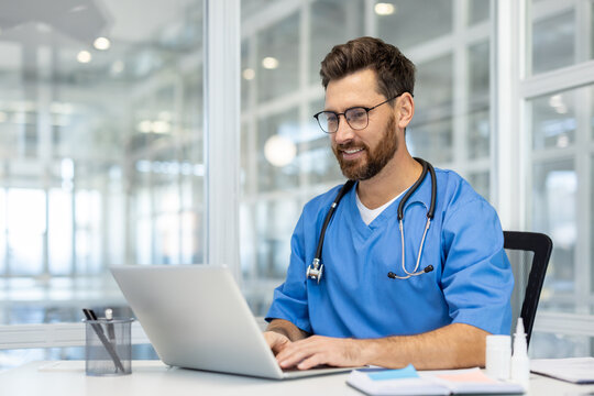 Smiling healthcare professional in scrubs using laptop in modern office setting with stethoscope. Represents productivity, professionalism, healthcare, technology integration in medical field.