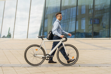 Fototapeta premium A Man Walks Alongside His Bicycle Near a Modern Glass Building on a Sunny Day