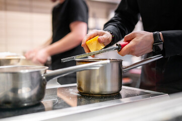 A close-up of hands grating cheese over a pot on a stove, with another person cooking in the background. The kitchen scene is warm and inviting, showcasing culinary preparation and teamwork