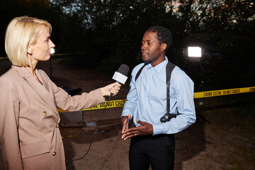 Waist up portrait of female reporter interviewing police officer speaking to microphone at crime...