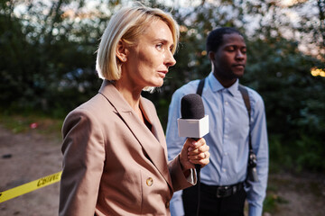 Full length portrait of woman speaking to microphone while recording news at crime scene with...