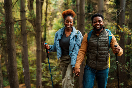 Happy black couple hiking in a lush green forest, holding hands and smiling. They are dressed in casual outdoor clothing, with hiking poles in hand. - Powered by Adobe