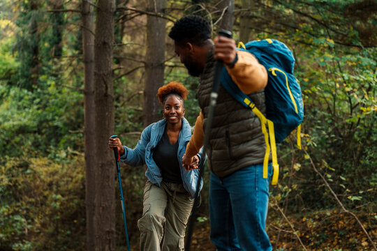 A black couple hiking in a forest, using trekking poles, with backpacks. men helping by giving a hand of help over a rocky path.