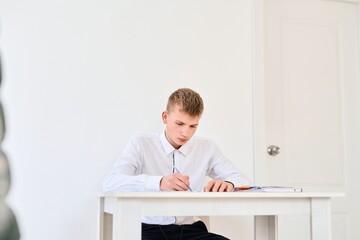 Obraz premium Portrait of a high school student in a white uniform doing homework, sitting at a table with textbooks, writing in a notebook