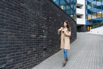 Young confident businesswoman standing outdoor of office center building on the street in a business suit and using smart phone, typing messages, texting. Design professional occupation female worker