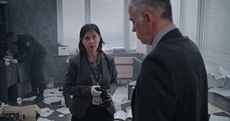 Office robbery case: Crime investigation team at crime scene. Female criminalist talks to male detective, then takes photos of clues and evidence. African American policeman works in the background.