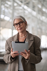 Elderly businesswoman standing confidently outside an office building, engaging with a tablet...