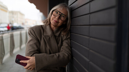 Elderly businesswoman standing outside an office, engaging with her smartphone on a cool day in the city