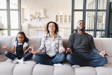 Serene african-american family of three, parents with small child kid daughter meditating together...