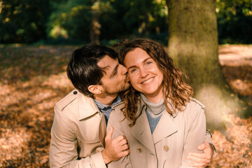 Happy smiling beautiful romantic couple in trench coats in the autumn park, a man kisses a woman