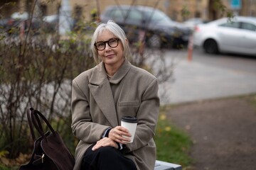 An older woman enjoys a warm drink while seated on a park bench during a cool autumn afternoon