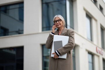 Senior businesswoman standing outside her office, discussing on the phone while holding a laptop...