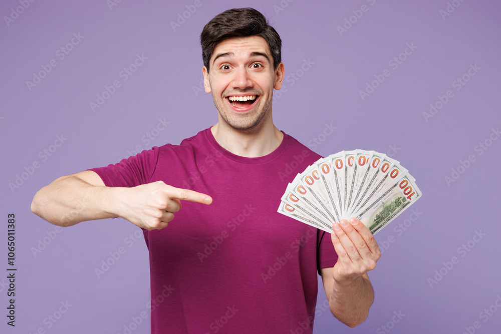 Wall mural Young Caucasian man he wear violet t-shirt casual clothes hold in hand point finger on fan of cash money in dollar banknotes isolated on plain pastel light purple background studio. Lifestyle concept.