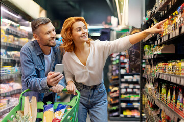 Couple doing grocery shopping together, man using cellphone, woman pointing at shelf, choosing food products in supermarket