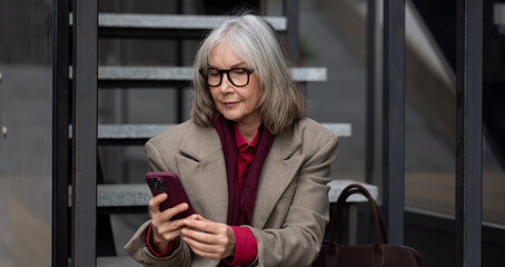 Fototapeta premium Middle-aged businesswoman in a trendy urban setting checking her smartphone while sitting on steps on a sunny day