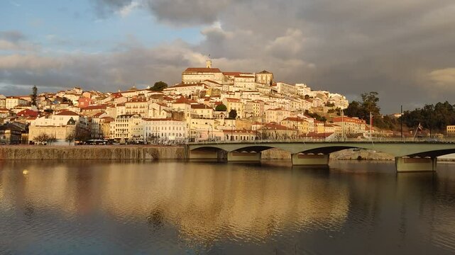 beautiful cityscape of Coimbra, Portugal, during golden hour. The warm sunlight bathes the historic buildings on the hillside, casting a rich, golden glow over their facades. The iconic university bui