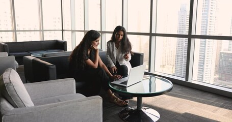 Two diverse young female entrepreneurs discussing online startup presentation on laptop, meeting for professional networking, talking on couch in co-working space interior with city view