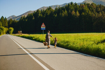 Mom and daughter walk along the road by the hand against the background of the forest and mountains. Mom and a girl in dresses. There is a picnic basket in his hands. Italian alps, countryside, tranqu