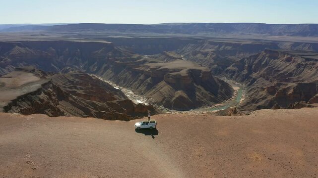 Aerial: Slow panning drone shot of one man standing on the roof of a white pick up truck near the edge of Fish river canyon in Namibia, Africa