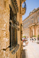 An antique beautiful metal grate on narrow medieval street with lanterns and beautiful flower trees in the old town of  Mdina on a sunny day, the silent city, ancient capital of Malta, Europe.