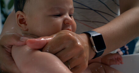 Hands washing baby’s arm during bath, protective and caring gesture, baby looks to the side,...