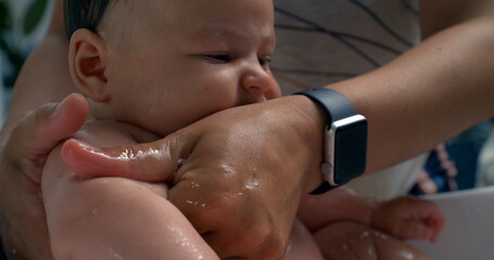 Hands washing baby’s arm during bath, protective and caring gesture, baby looks to the side,...