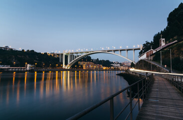 Obraz premium Arrabida bridge with car light trails reflecting on douro river at blue hour with illuminated cityscape and wooden walkway in Porto, Portugal