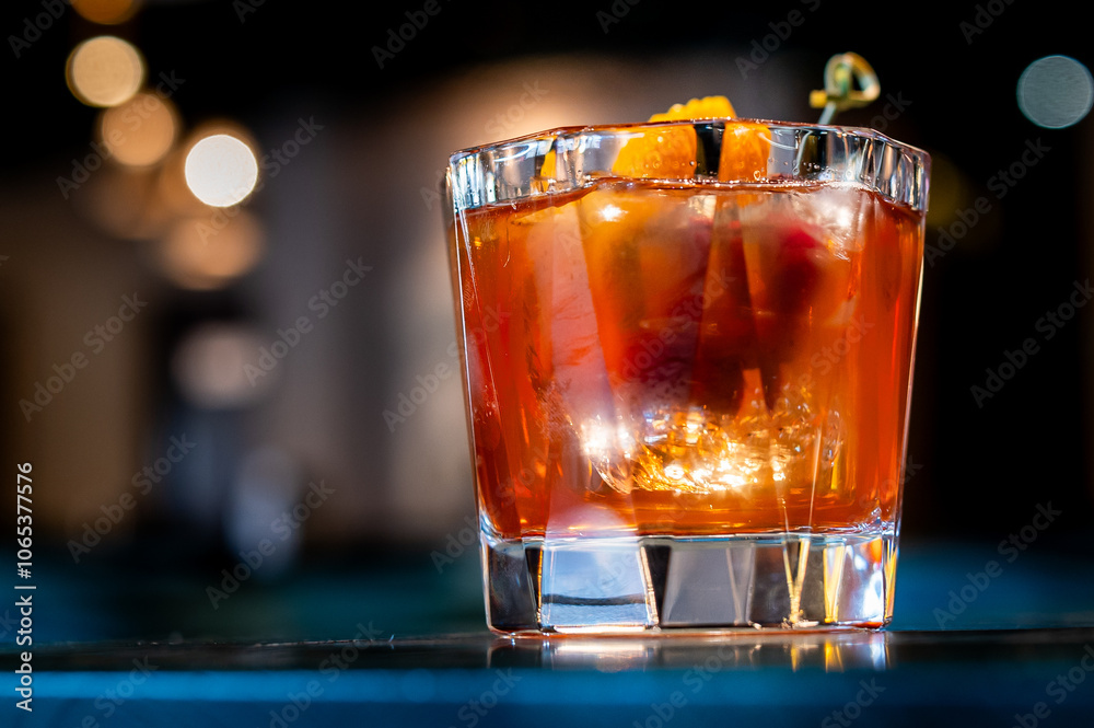 Wall mural A close-up of a vibrant, amber-colored cocktail in a short glass, garnished with an orange slice and cherry, set against a blurred bar background. Perfect for themes of nightlife