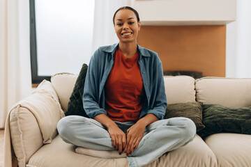 Fototapeta premium Portrait of smiling, happy African American woman sitting on comfortable sofa in lotus position
